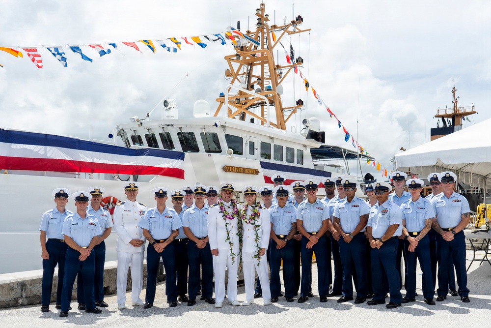 A group of uniformed Coast Guard personnel pose for a formal group photograph in front of a white Coast Guard vessel named 'Oliver Henry'. The occasion appears to be a commissioning or decommissioning ceremony, evidenced by the colorful signal flags (dress ship bunting) strung across the vessel and two central figures wearing traditional Hawaiian lei garlands. The setting is a dock or pier in what appears to be a tropical location.
