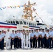 A group of uniformed Coast Guard personnel pose for a formal group photograph in front of a white Coast Guard vessel named 'Oliver Henry'. The occasion appears to be a commissioning or decommissioning ceremony, evidenced by the colorful signal flags (dress ship bunting) strung across the vessel and two central figures wearing traditional Hawaiian lei garlands. The setting is a dock or pier in what appears to be a tropical location.