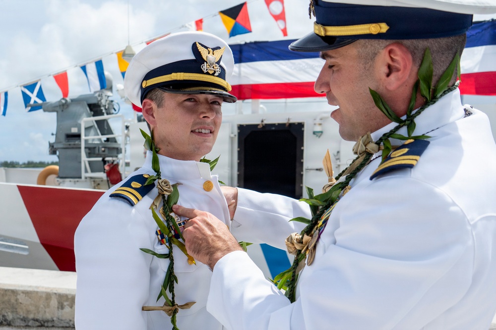 Two U.S. Coast Guard officers in white dress uniforms are participating in what appears to be a formal change-of-command or commissioning ceremony. One officer is pinning or adjusting a medal/insignia on the other's uniform while both wear traditional Hawaiian lei garlands, suggesting the event is taking place in Hawaii. Colorful nautical signal flags are visible in the background aboard what appears to be a Coast Guard vessel.