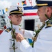 Two U.S. Coast Guard officers in white dress uniforms are participating in what appears to be a formal change-of-command or commissioning ceremony. One officer is pinning or adjusting a medal/insignia on the other's uniform while both wear traditional Hawaiian lei garlands, suggesting the event is taking place in Hawaii. Colorful nautical signal flags are visible in the background aboard what appears to be a Coast Guard vessel.