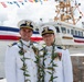 Two U.S. Coast Guard officers in dress white uniforms pose for a photo at what appears to be a ship commissioning ceremony. They are wearing traditional Hawaiian lei garlands and display multiple service ribbons and medals on their uniforms. The vessel visible behind them is named 'Oliver Henry,' decorated with colorful signal flags for the occasion.