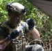A military medic or corpsman in full tactical gear is preparing a medical syringe in an outdoor jungle or forested environment. The individual is wearing a combat helmet with mounted accessories, plate carrier vest with medical supplies, black tactical gloves, and aviator sunglasses. The scene appears to depict a field medical training exercise or real-world casualty care scenario.