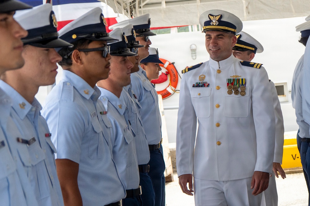 A senior officer in a white dress uniform with medals and gold insignia is inspecting or reviewing a line of personnel dressed in light blue uniforms and dark-brimmed caps. The setting appears to be a maritime or coast guard facility, with a white vessel visible in the background. The officer is smiling as he walks along the formation.