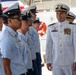 A senior officer in a white dress uniform with medals and gold insignia is inspecting or reviewing a line of personnel dressed in light blue uniforms and dark-brimmed caps. The setting appears to be a maritime or coast guard facility, with a white vessel visible in the background. The officer is smiling as he walks along the formation.