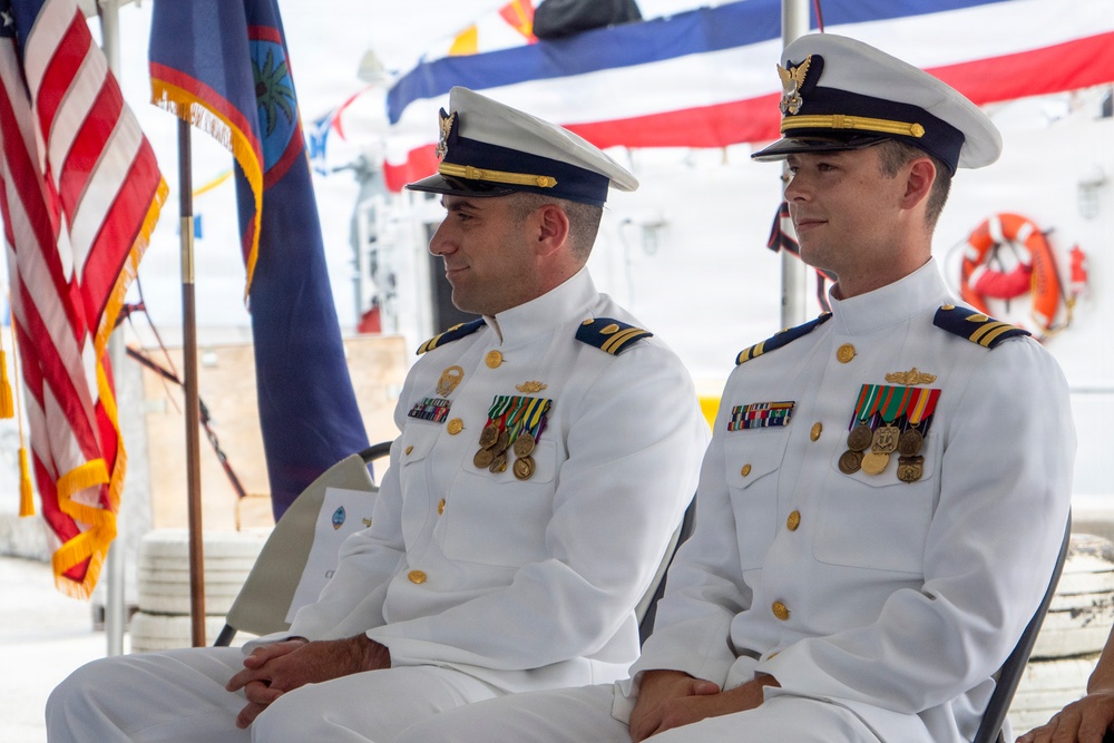Two U.S. Coast Guard officers in dress white uniforms are seated at what appears to be a formal military ceremony. Both officers wear service ribbons, medals, and rank insignia on their uniforms, suggesting they hold officer-level positions. The setting includes multiple flags, including the U.S. flag and what appears to be the flag of Guam, indicating this ceremony may be taking place in or near Guam.