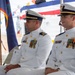 Two U.S. Coast Guard officers in dress white uniforms are seated at what appears to be a formal military ceremony. Both officers wear service ribbons, medals, and rank insignia on their uniforms, suggesting they hold officer-level positions. The setting includes multiple flags, including the U.S. flag and what appears to be the flag of Guam, indicating this ceremony may be taking place in or near Guam.