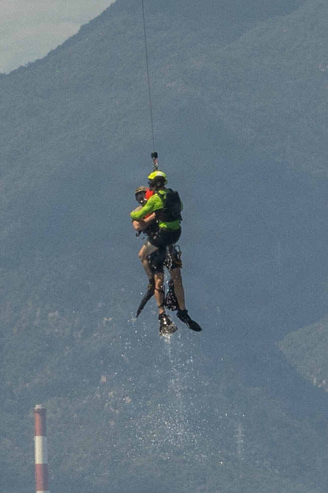 A helicopter rescue operation is underway, showing a rescuer in a bright yellow-green helmet and vest hoisting a person via a cable hoist line. Water droplets are visibly falling from the rescued individual, suggesting they were recently extracted from water. The scene is set against a mountainous backdrop with a red-and-white striped marker pole visible in the lower left.