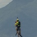 A helicopter rescue operation is underway, showing a rescuer in a bright yellow-green helmet and vest hoisting a person via a cable hoist line. Water droplets are visibly falling from the rescued individual, suggesting they were recently extracted from water. The scene is set against a mountainous backdrop with a red-and-white striped marker pole visible in the lower left.