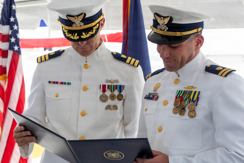 Two U.S. Coast Guard officers in white dress uniforms are reviewing an official document or certificate during what appears to be a formal military ceremony. Both officers wear captain-rank insignia and display multiple service medals and ribbons. An American flag and additional ceremonial flags are visible in the background.