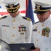 Two U.S. Coast Guard officers in white dress uniforms are reviewing an official document or certificate during what appears to be a formal military ceremony. Both officers wear captain-rank insignia and display multiple service medals and ribbons. An American flag and additional ceremonial flags are visible in the background.