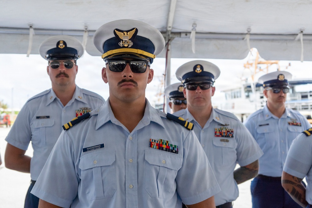 A group of U.S. Coast Guard personnel in dress uniform are standing in formation, likely during a formal ceremony or official event. The central figure, identified by a name tag reading 'WASSERMAN,' wears an officer's cover with gold insignia and displays multiple service ribbons. The setting appears to be an outdoor waterfront location under a white tent canopy.