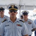 A group of U.S. Coast Guard personnel in dress uniform are standing in formation, likely during a formal ceremony or official event. The central figure, identified by a name tag reading 'WASSERMAN,' wears an officer's cover with gold insignia and displays multiple service ribbons. The setting appears to be an outdoor waterfront location under a white tent canopy.