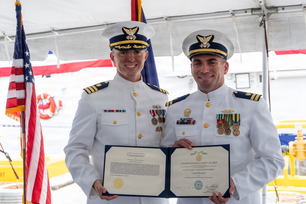 Two United States Coast Guard officers in white dress uniforms are posing together while holding an open certificate folder displaying a 'Coast Guard Commendation Medal' award document. The ceremony appears to be taking place outdoors under a tent, likely aboard or near a Coast Guard vessel. Both officers are smiling and wearing service medals and ribbons on their uniforms.