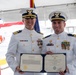 Two United States Coast Guard officers in white dress uniforms are posing together while holding an open certificate folder displaying a 'Coast Guard Commendation Medal' award document. The ceremony appears to be taking place outdoors under a tent, likely aboard or near a Coast Guard vessel. Both officers are smiling and wearing service medals and ribbons on their uniforms.