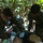 Two military personnel in a jungle/forest environment are providing medical treatment to a casualty on the ground. One individual is wearing full camouflage uniform with a tactical backpack, while the other is in a olive green t-shirt with camouflage trousers. This appears to be either a field medical training exercise or an actual combat casualty care scenario.