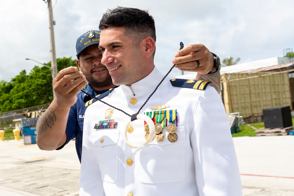 A U.S. Coast Guard officer in white dress uniform is having a traditional lei or necklace placed around his neck by an enlisted Coast Guard member wearing a blue utility uniform and a 'U.S. Coast Guard Base Guam' cap. The officer displays multiple service medals, ribbons, and gold insignia on his dress whites, suggesting a formal ceremony or recognition event. The setting appears to be an outdoor base facility, likely in Guam based on the tropical vegetation and cap insignia.