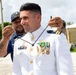 A U.S. Coast Guard officer in white dress uniform is having a traditional lei or necklace placed around his neck by an enlisted Coast Guard member wearing a blue utility uniform and a 'U.S. Coast Guard Base Guam' cap. The officer displays multiple service medals, ribbons, and gold insignia on his dress whites, suggesting a formal ceremony or recognition event. The setting appears to be an outdoor base facility, likely in Guam based on the tropical vegetation and cap insignia.
