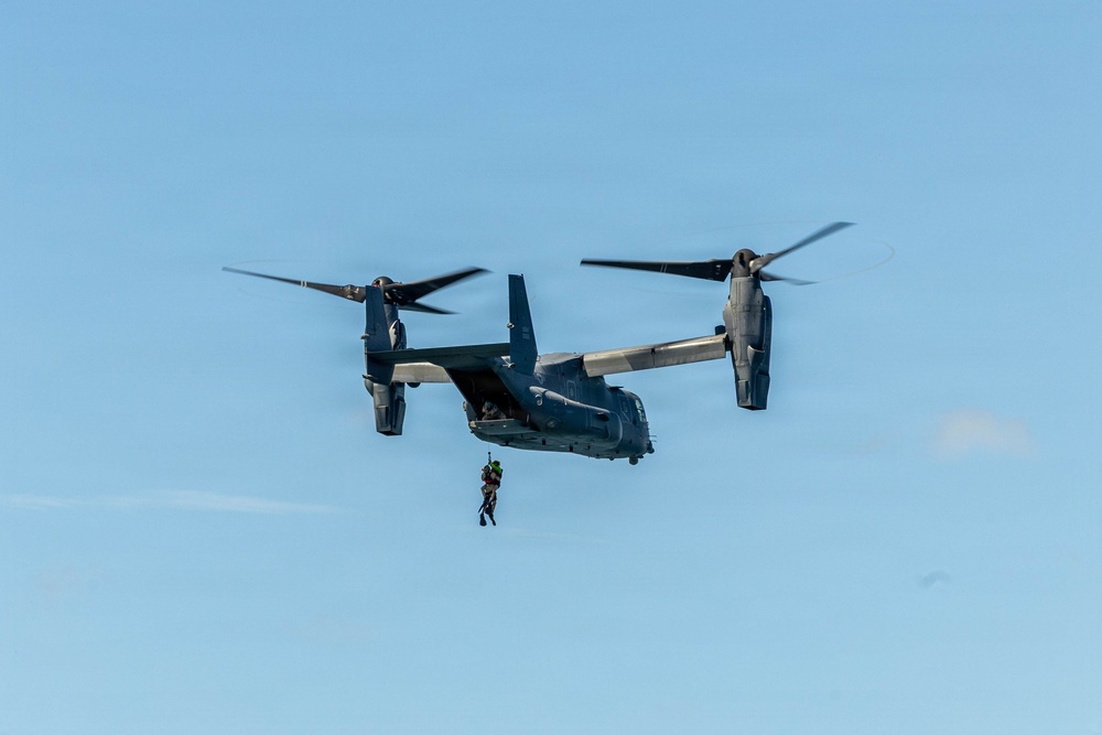 A military tiltrotor aircraft, identifiable as a Bell Boeing V-22 Osprey, is hovering in flight against a clear blue sky. A person in tactical gear is suspended below the aircraft on a hoist or rope, suggesting a rescue or insertion operation is underway. The aircraft's rotors are tilted upward in helicopter mode, consistent with a hover or low-speed flight configuration.