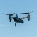 A military tiltrotor aircraft, identifiable as a Bell Boeing V-22 Osprey, is hovering in flight against a clear blue sky. A person in tactical gear is suspended below the aircraft on a hoist or rope, suggesting a rescue or insertion operation is underway. The aircraft's rotors are tilted upward in helicopter mode, consistent with a hover or low-speed flight configuration.