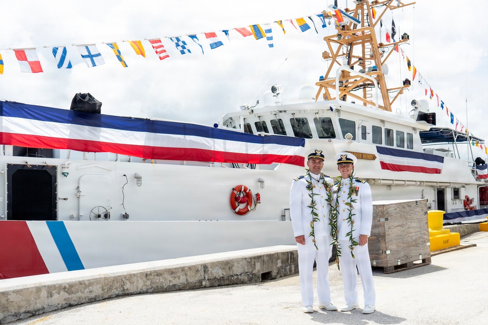 Two naval/coast guard officers in white dress uniforms are posing in front of a large white vessel decorated with international signal flags and a large ceremonial banner in red, white, and blue stripes. Both individuals are wearing traditional Hawaiian lei garlands, suggesting this is a commissioning or decommissioning ceremony. The setting appears to be a dock or pier in a tropical location.