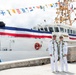 Two naval/coast guard officers in white dress uniforms are posing in front of a large white vessel decorated with international signal flags and a large ceremonial banner in red, white, and blue stripes. Both individuals are wearing traditional Hawaiian lei garlands, suggesting this is a commissioning or decommissioning ceremony. The setting appears to be a dock or pier in a tropical location.