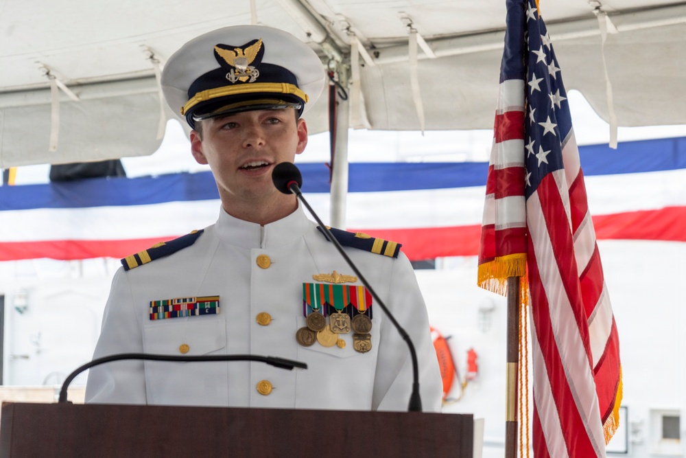 A U.S. Coast Guard officer in dress whites is speaking at a podium during a formal military ceremony. The officer is wearing multiple service ribbons, medals, and rank insignia consistent with a lieutenant commander or commander grade. An American flag and red-white-blue bunting are visible in the background, suggesting a commissioning, change of command, or similar official event.