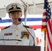 A U.S. Coast Guard officer in dress whites is speaking at a podium during a formal military ceremony. The officer is wearing multiple service ribbons, medals, and rank insignia consistent with a lieutenant commander or commander grade. An American flag and red-white-blue bunting are visible in the background, suggesting a commissioning, change of command, or similar official event.