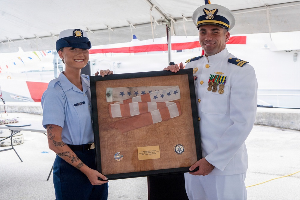 Two U.S. Coast Guard personnel are posing together holding a framed shadow box containing a folded American flag and two unit coins with a commemorative plaque. The officer on the right is in dress whites with medals and rank insignia, while the enlisted member on the left is in the standard blue uniform. This appears to be a military ceremony, likely a change of command or retirement event.