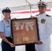 Two U.S. Coast Guard personnel are posing together holding a framed shadow box containing a folded American flag and two unit coins with a commemorative plaque. The officer on the right is in dress whites with medals and rank insignia, while the enlisted member on the left is in the standard blue uniform. This appears to be a military ceremony, likely a change of command or retirement event.