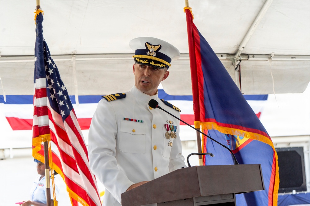 A uniformed military officer, appearing to be a U.S. Coast Guard Captain or Commander, is delivering a speech at a formal ceremony from a wooden podium. The setting is an outdoor event under a white tent, flanked by the U.S. flag and what appears to be the flag of Guam. The officer is wearing a white dress uniform adorned with multiple medals and ribbons.