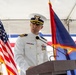 A uniformed military officer, appearing to be a U.S. Coast Guard Captain or Commander, is delivering a speech at a formal ceremony from a wooden podium. The setting is an outdoor event under a white tent, flanked by the U.S. flag and what appears to be the flag of Guam. The officer is wearing a white dress uniform adorned with multiple medals and ribbons.