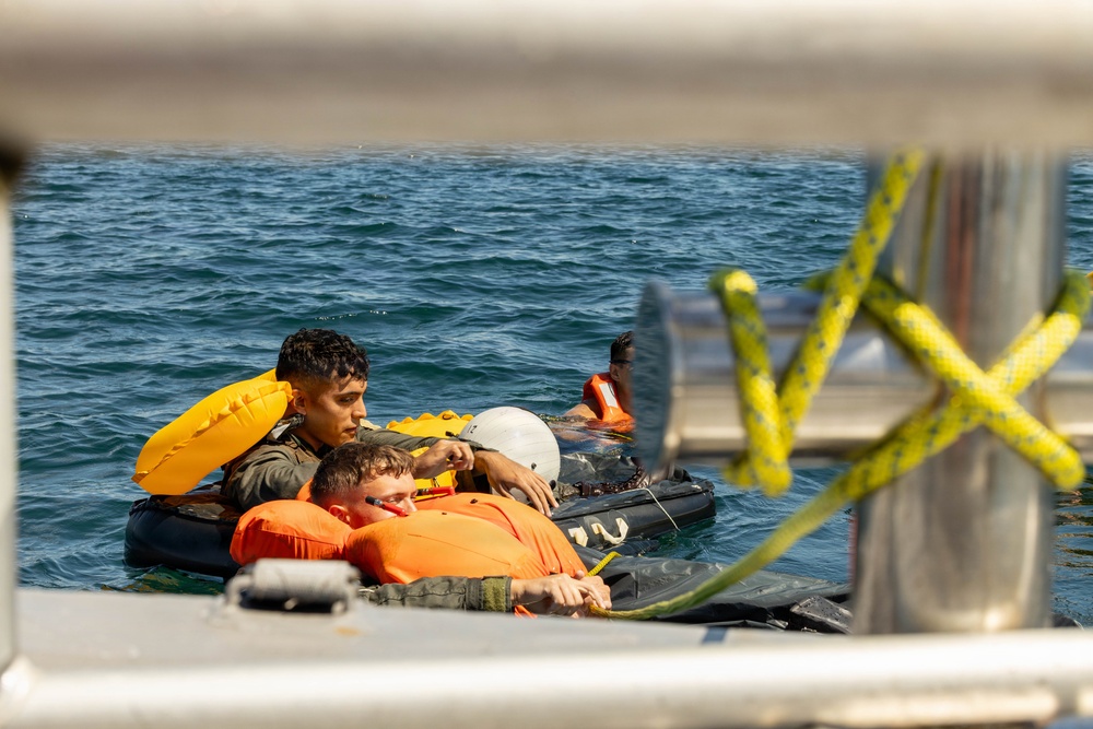 The image shows multiple individuals in the water clinging to what appears to be a small inflatable raft or flotation device near a vessel. The people are wearing orange life vests and survival gear, suggesting a water survival training exercise or rescue scenario. The photo is taken from aboard a nearby vessel, with yellow safety ropes visible in the foreground.