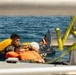 The image shows multiple individuals in the water clinging to what appears to be a small inflatable raft or flotation device near a vessel. The people are wearing orange life vests and survival gear, suggesting a water survival training exercise or rescue scenario. The photo is taken from aboard a nearby vessel, with yellow safety ropes visible in the foreground.