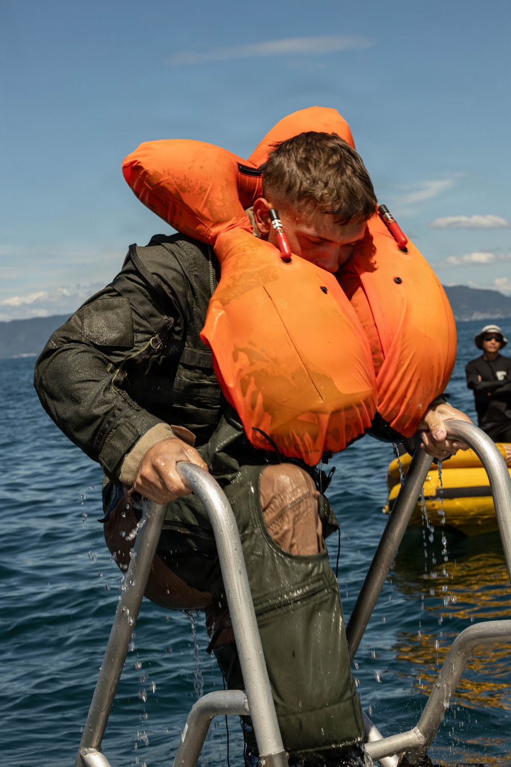 A person wearing an inflated orange life vest and wet olive/green military-style clothing is climbing a metal ladder out of open water. Water is visibly dripping from their clothing and the ladder, suggesting they have just emerged from the sea. A second individual in dark clothing and sunglasses is visible in the background near a yellow inflatable vessel.