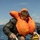 U.S. Marines, JMSDF Sailors reinforce maritime readiness during Rescue Flag 26-1 A person wearing an inflated orange life vest and wet olive/green military-style clothing is climbing a metal ladder out of open water. Water is visibly dripping from their clothing and the ladder, suggesting they have just emerged from the sea. A second individual in dark clothing and sunglasses is visible in the background near a yellow inflatable vessel.