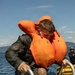 A person wearing an inflated orange life vest and wet olive/green military-style clothing is climbing a metal ladder out of open water. Water is visibly dripping from their clothing and the ladder, suggesting they have just emerged from the sea. A second individual in dark clothing and sunglasses is visible in the background near a yellow inflatable vessel.