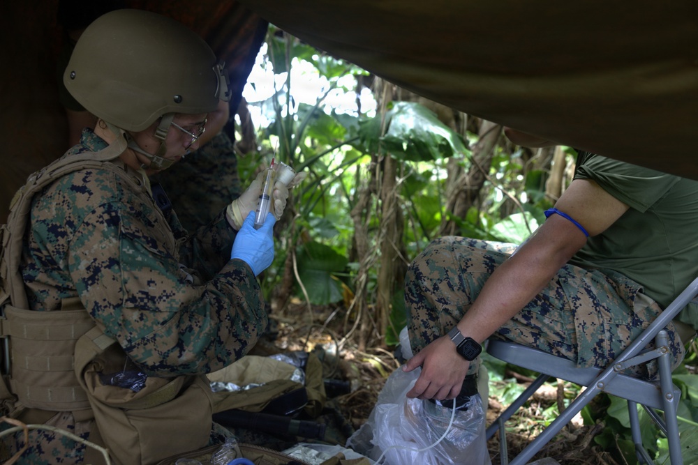 A military medic in full combat gear, including helmet and body armor, is preparing or administering a medical procedure using a syringe in a jungle field setting. A second service member in camouflage uniform is seated nearby, appearing to be the patient, with a blue tourniquet or band visible on their arm. The scene takes place under a makeshift shelter or tarp in a dense tropical environment.