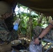 A military medic in full combat gear, including helmet and body armor, is preparing or administering a medical procedure using a syringe in a jungle field setting. A second service member in camouflage uniform is seated nearby, appearing to be the patient, with a blue tourniquet or band visible on their arm. The scene takes place under a makeshift shelter or tarp in a dense tropical environment.