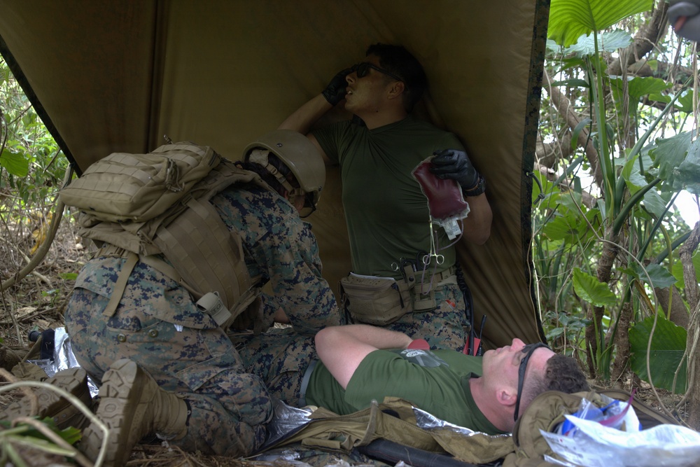 The image shows what appears to be a military medical training exercise in a jungle or forested environment. A person in USMC digital camouflage uniform with a tactical backpack is kneeling over a simulated casualty lying on a mylar emergency blanket, while a second individual in an olive drab t-shirt holds what appears to be a blood/IV bag and medical instruments. The scene is set under an olive-colored tarp shelter amid dense tropical vegetation.