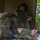 The image shows what appears to be a military medical training exercise in a jungle or forested environment. A person in USMC digital camouflage uniform with a tactical backpack is kneeling over a simulated casualty lying on a mylar emergency blanket, while a second individual in an olive drab t-shirt holds what appears to be a blood/IV bag and medical instruments. The scene is set under an olive-colored tarp shelter amid dense tropical vegetation.