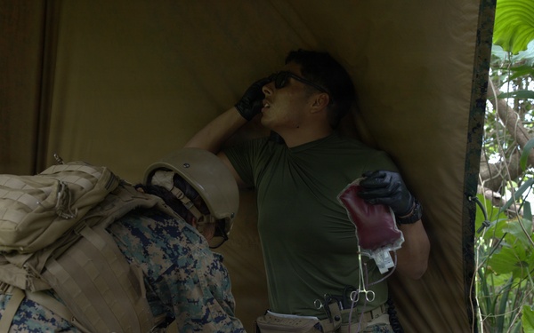 The image shows what appears to be a military medical training exercise in a jungle or forested environment. A person in USMC digital camouflage uniform with a tactical backpack is kneeling over a simulated casualty lying on a mylar emergency blanket, while a second individual in an olive drab t-shirt holds what appears to be a blood/IV bag and medical instruments. The scene is set under an olive-colored tarp shelter amid dense tropical vegetation.
