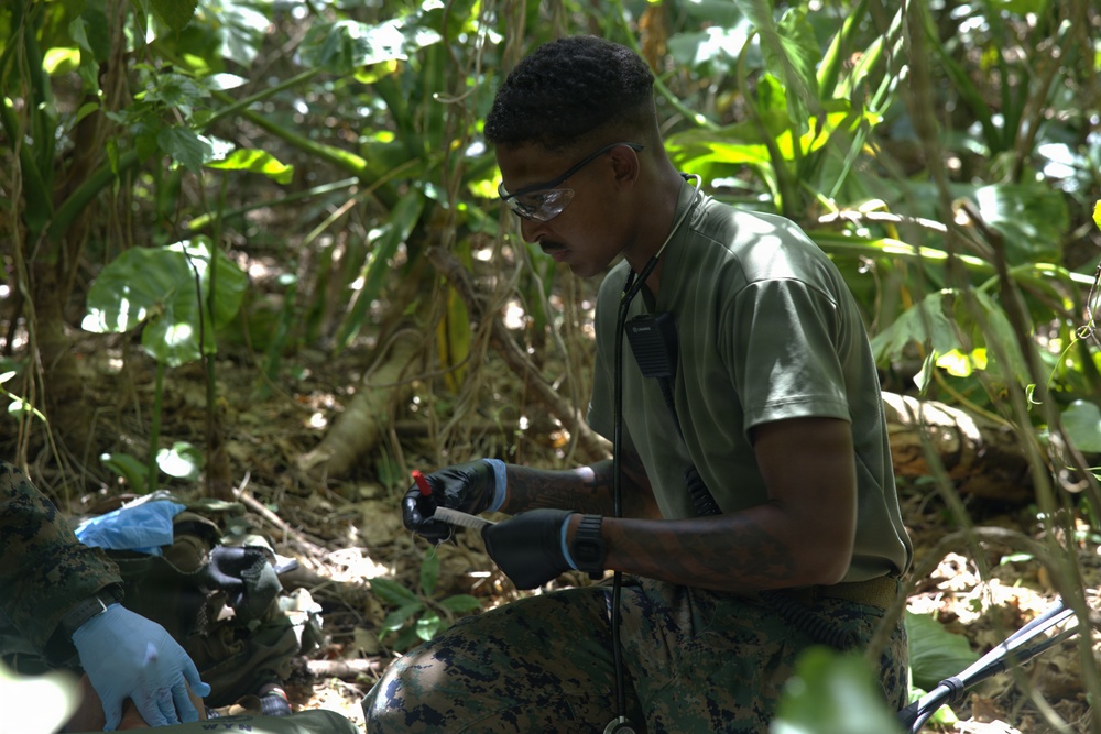 A military service member wearing olive drab clothing and MARPAT camouflage trousers is working in a jungle or dense vegetation environment. He is wearing safety glasses and black gloves while handling what appears to be a small object or tool, possibly conducting an EOD (Explosive Ordnance Disposal) or field medical/technical task. A second person wearing blue latex gloves is partially visible on the left side of the frame.