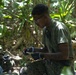 A military service member wearing olive drab clothing and MARPAT camouflage trousers is working in a jungle or dense vegetation environment. He is wearing safety glasses and black gloves while handling what appears to be a small object or tool, possibly conducting an EOD (Explosive Ordnance Disposal) or field medical/technical task. A second person wearing blue latex gloves is partially visible on the left side of the frame.