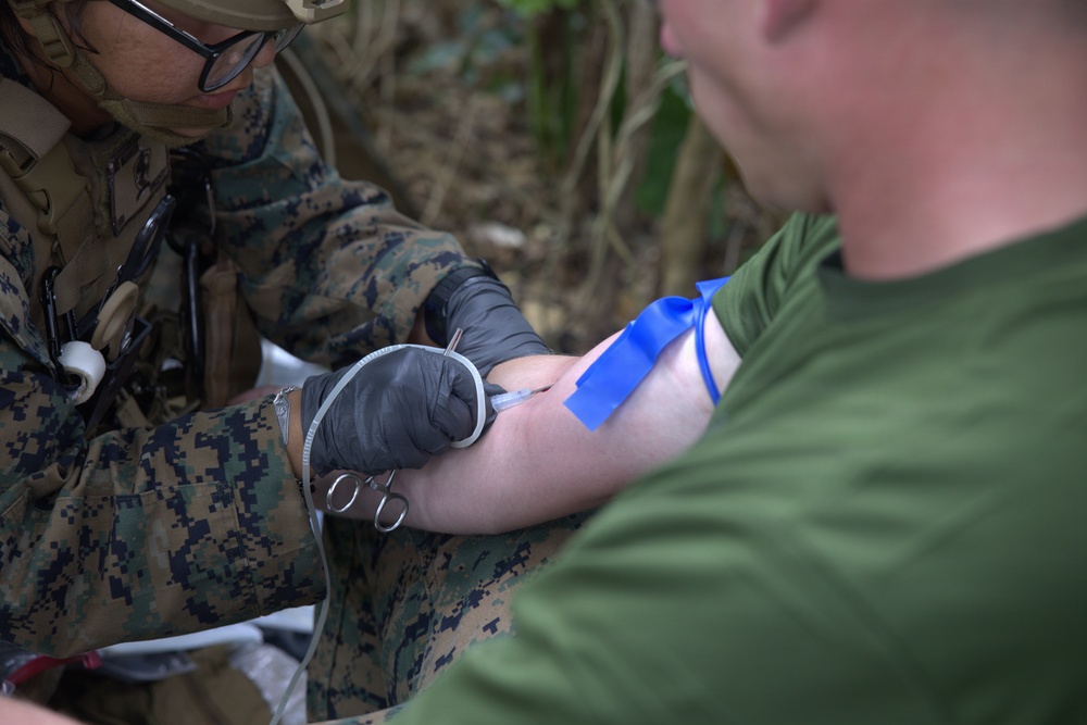 A military medic in MARPAT digital camouflage uniform is performing an intravenous (IV) insertion or blood draw on a patient wearing a green t-shirt. The medic is wearing black nitrile gloves and using a needle/catheter on the patient's arm, which has a blue tourniquet or tape applied above the insertion site. The setting appears to be an outdoor field environment, consistent with a tactical combat casualty care (TCCC) scenario.
