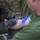 A military medic in MARPAT digital camouflage uniform is performing an intravenous (IV) insertion or blood draw on a patient wearing a green t-shirt. The medic is wearing black nitrile gloves and using a needle/catheter on the patient's arm, which has a blue tourniquet or tape applied above the insertion site. The setting appears to be an outdoor field environment, consistent with a tactical combat casualty care (TCCC) scenario.