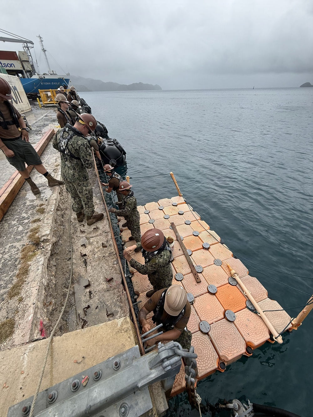 A group of military personnel, appearing to be U.S. Navy sailors wearing camouflage uniforms and hard hats, are assembling or securing a modular floating dock/pontoon system at a pier. The orange interlocking float modules are being connected to a concrete quay wall using chains and ropes. A vessel identified as 'KYOWA EAGLE' is visible in the background at the dock.