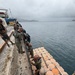 A group of military personnel, appearing to be U.S. Navy sailors wearing camouflage uniforms and hard hats, are assembling or securing a modular floating dock/pontoon system at a pier. The orange interlocking float modules are being connected to a concrete quay wall using chains and ropes. A vessel identified as 'KYOWA EAGLE' is visible in the background at the dock.