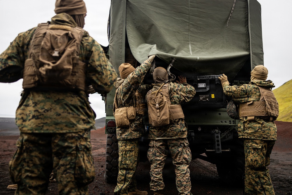 Four military personnel in MARPAT (Marine pattern) camouflage uniforms are gathered around the rear of a military tactical vehicle with an olive-drab canvas cover. Three soldiers are actively working at or looking into the back of the vehicle while a fourth observes from a short distance. The terrain appears dark and volcanic, suggesting a cold-weather training environment.
