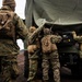 Four military personnel in MARPAT (Marine pattern) camouflage uniforms are gathered around the rear of a military tactical vehicle with an olive-drab canvas cover. Three soldiers are actively working at or looking into the back of the vehicle while a fourth observes from a short distance. The terrain appears dark and volcanic, suggesting a cold-weather training environment.