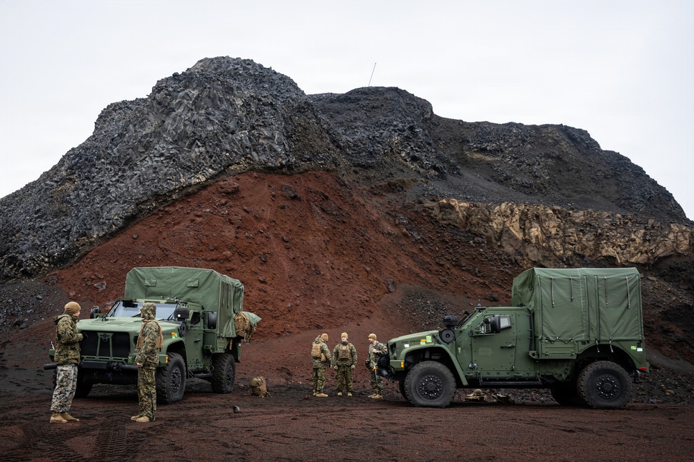 A group of military personnel in camouflage uniforms are gathered between two olive-green tactical military vehicles, identified as Joint Light Tactical Vehicles (JLTVs), in a rugged volcanic or rocky terrain. The landscape features dramatic multi-colored geological formations with dark basaltic rock, red iron-rich soil, and lighter sedimentary layers in the background. The overcast sky and barren terrain suggest a remote, possibly arctic or volcanic environment.